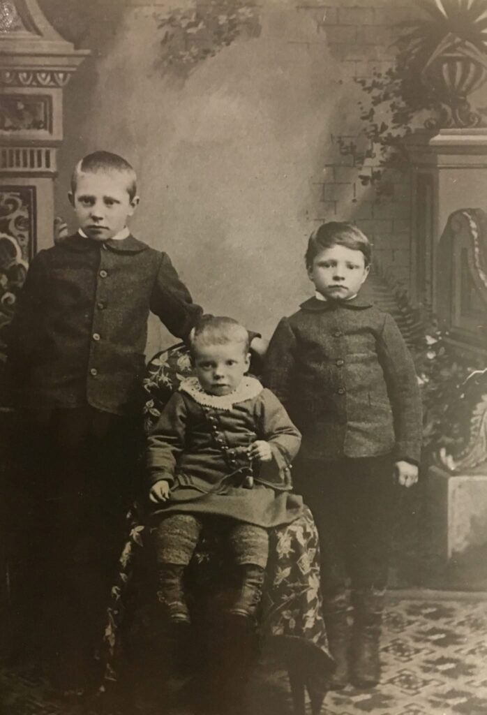 Three young children in old-fashioned clothing pose for a vintage black and white studio portrait; Baldur is on the left, Gudmundur is seated in the center, and Jakob on the right.