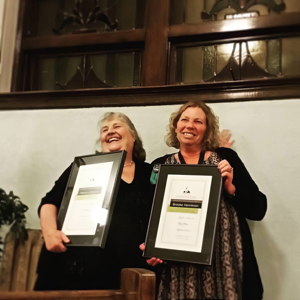 Two smiling women stand side by side, each holding a framed certificate. They are indoors in front of a wall, appearing happy and proud, at an awards ceremony.