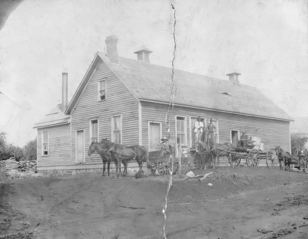 A black-and-white photo of a wooden house with people on the porch. Two horse-drawn carriages are parked in front on rough, unpaved ground—a rural scene reminiscent of the early 20th century, possibly before the local creamery closes in 1972.