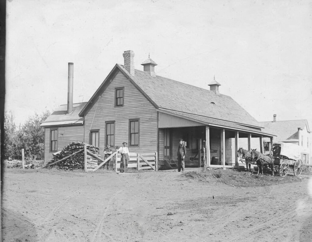 A black-and-white photo of the Creamery, wood piles, and a chimney. Three people stand outside the Creamery, and a horse-drawn carriage is on the right near a fence and trees.