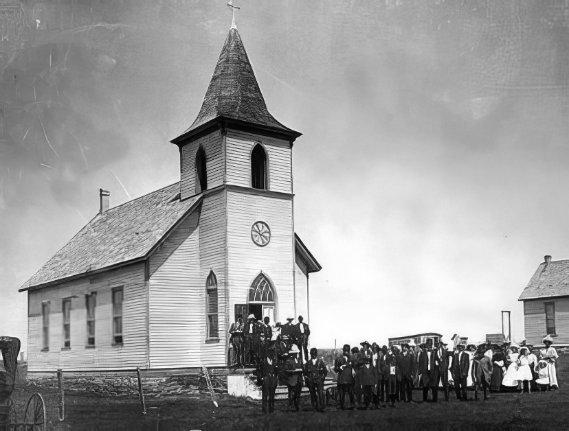 A black-and-white photo from between 1905 and 1907 shows a group gathered in front of a white wooden Lutheran Church with a tall steeple. Men, women, and children stand in formal attire on the grass and church steps.